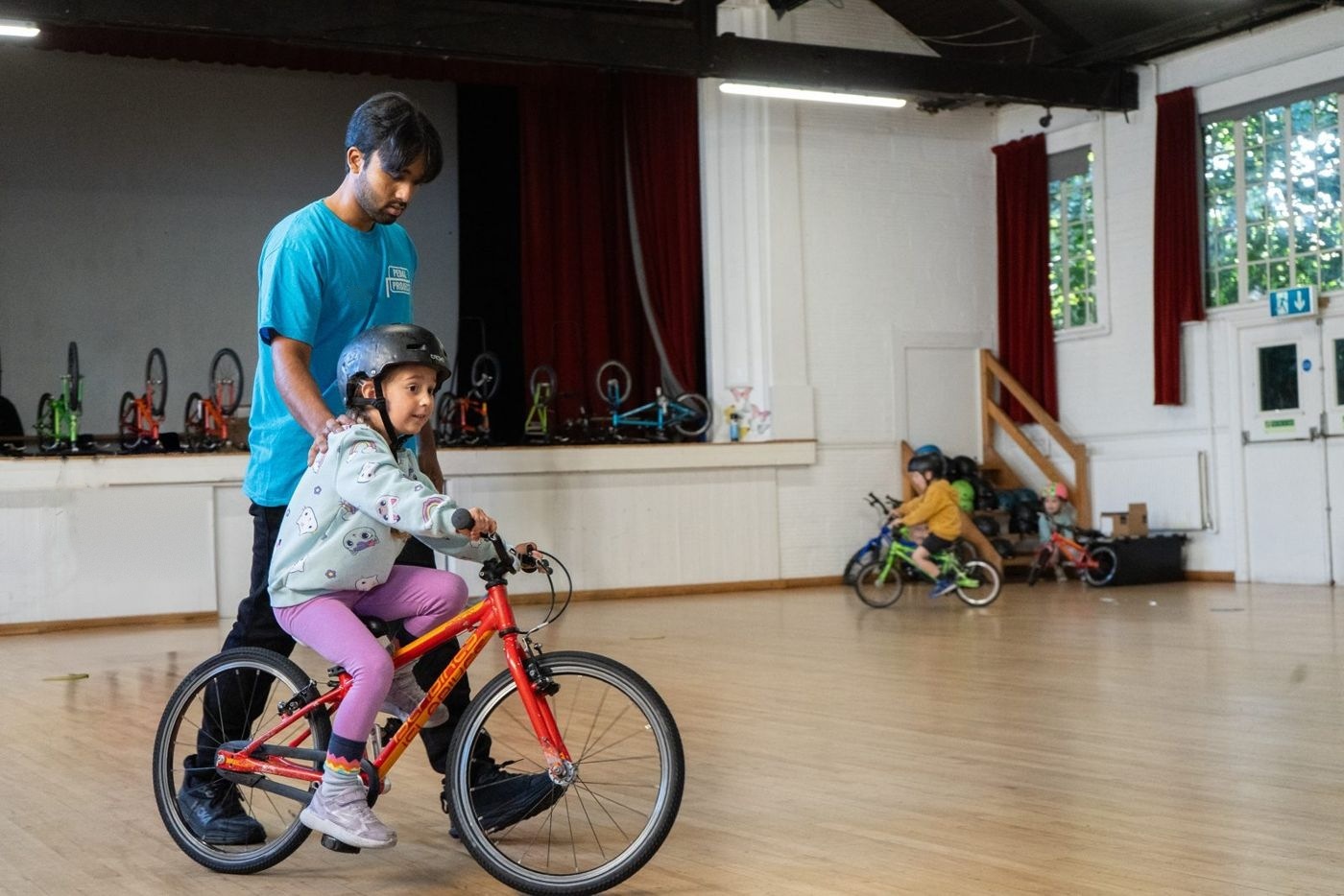 An instructor steadying a girl on an orange bike in a hall